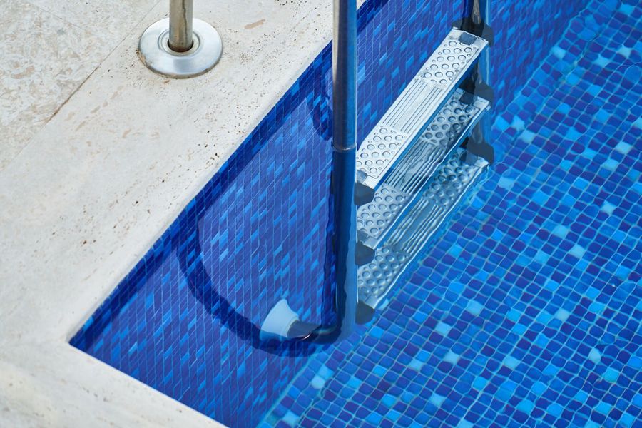 Pool ladder entering blue tiled water, with a metal handrail and white deck edge